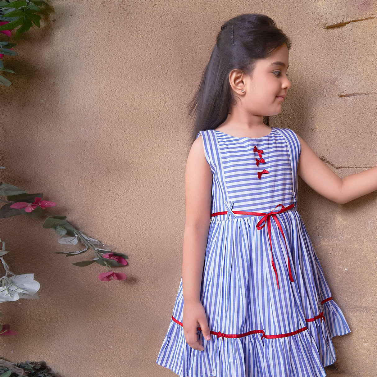 A little girl in sleeveless striped cotton dress with satin bows on hem ruffle and bodice and satin belt at wait