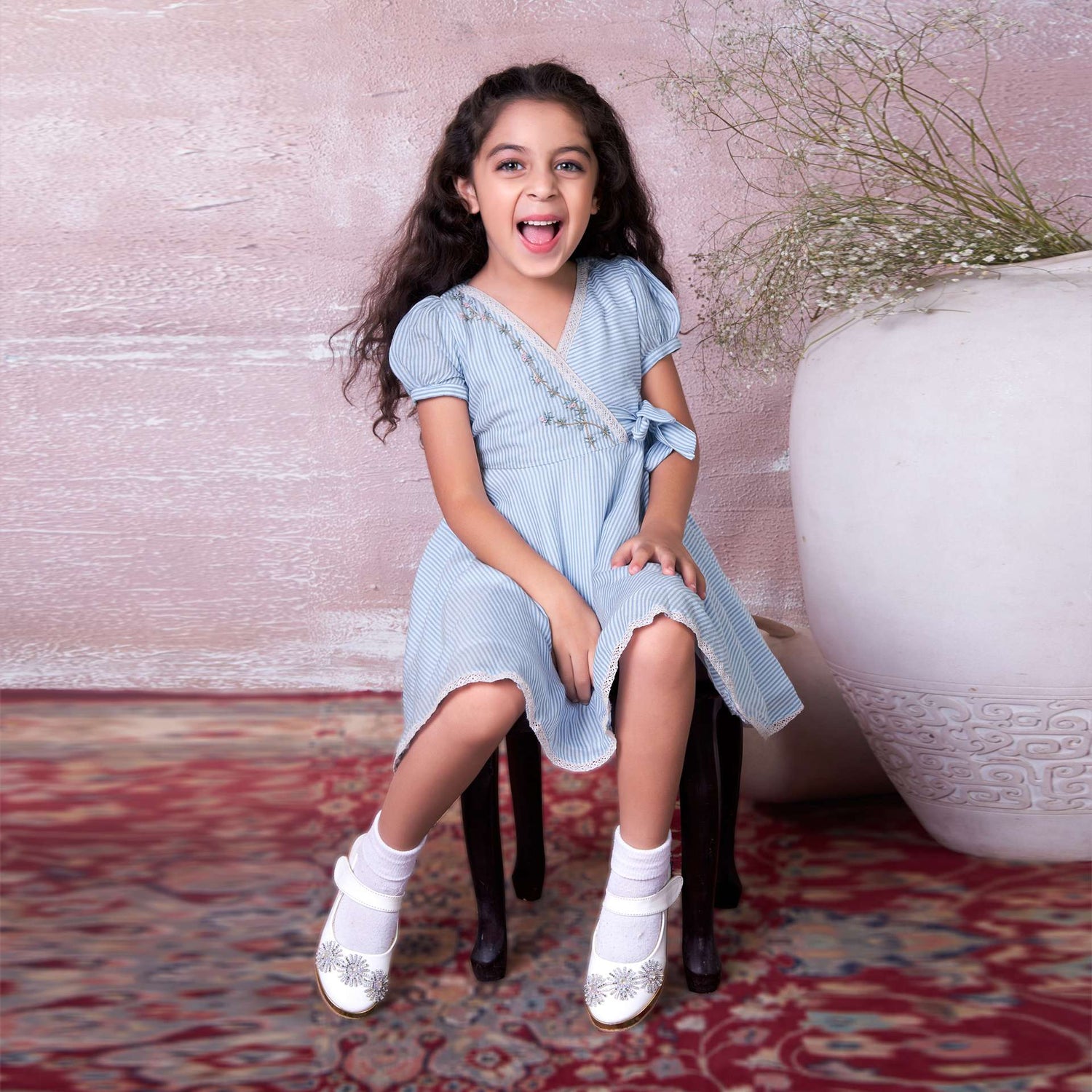 A girl seated on a stool wearing blue striped muslin cross over dress with crochet lace trims and hand embroidered bodice