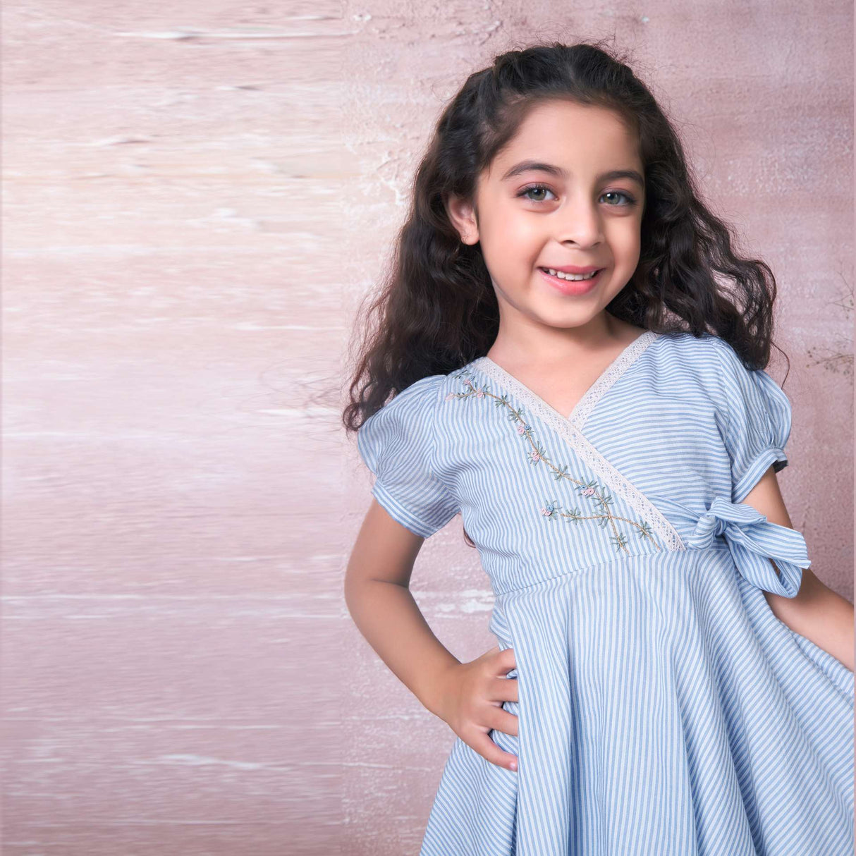 A girl smiling away wearing blue striped muslin dress with crossover bodice and hand embroidery