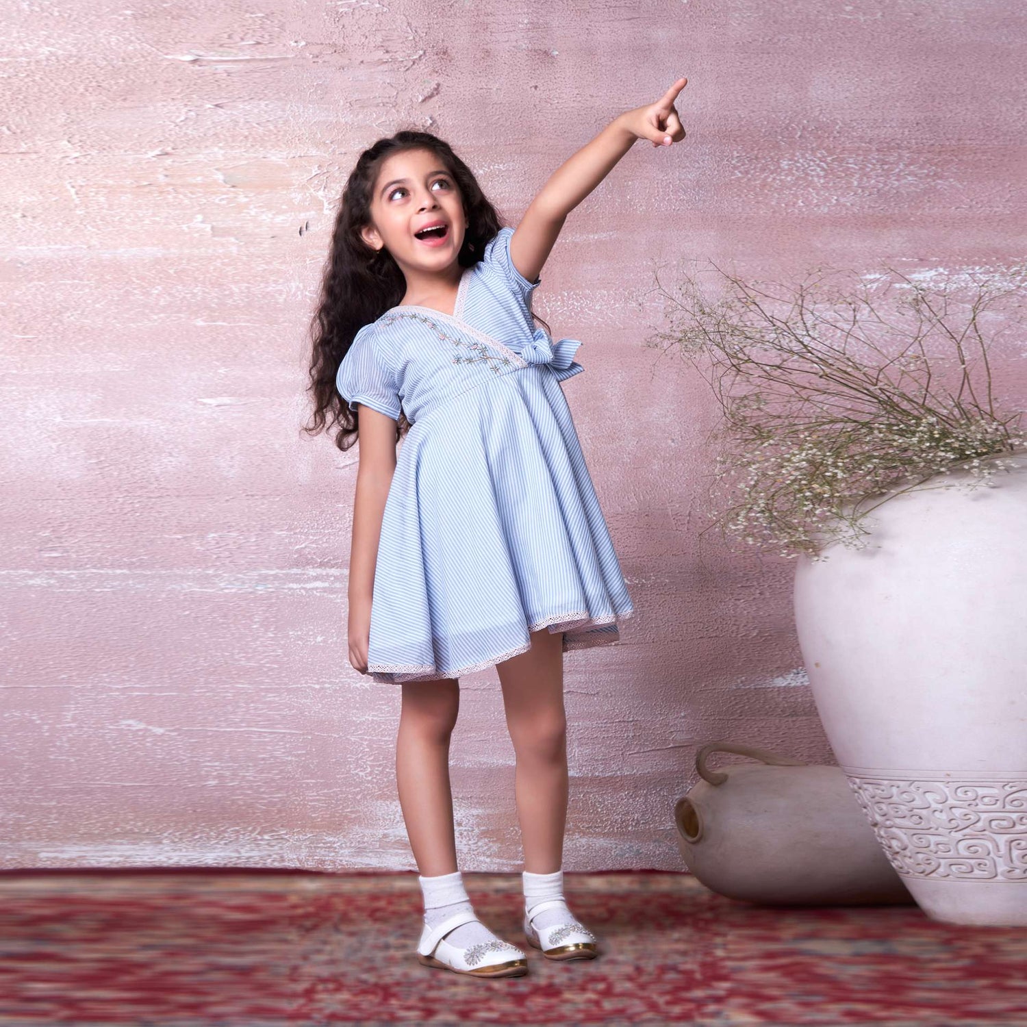 A girl in striped dress with crochet-style lace trim, intricate floral hand-embroidery, faux cross-over and side bow