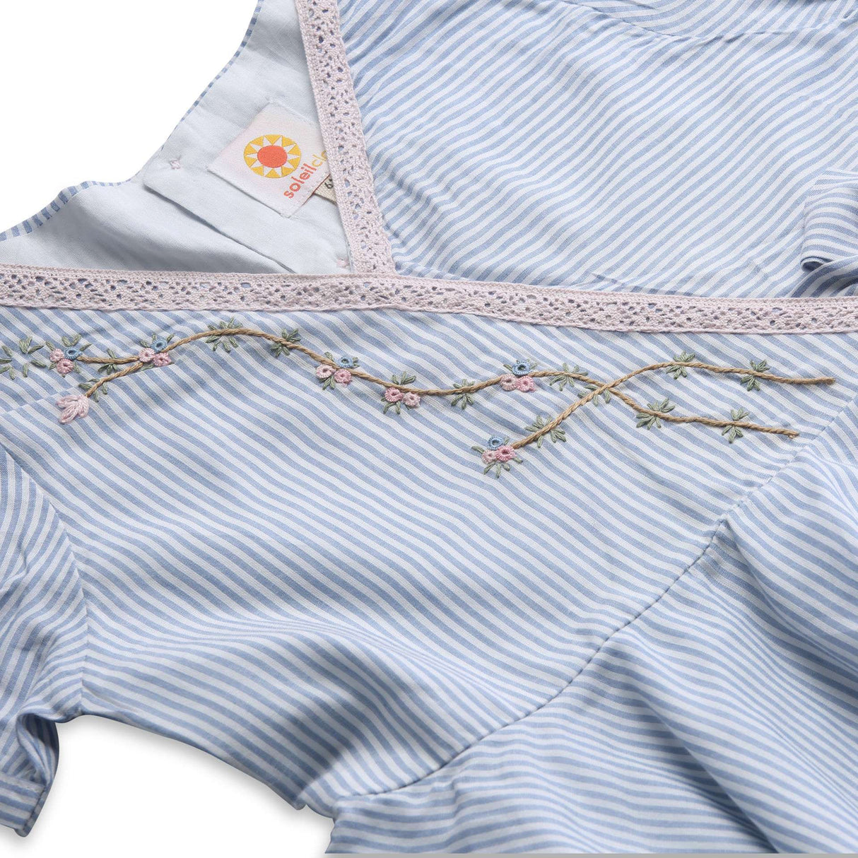 Flatlay of blue striped cotton dress with delicate crochet-style lace trim, floral hand-embroidery and faux cross-over