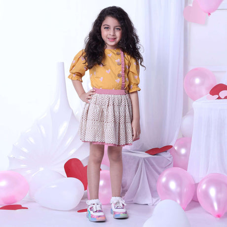 A girl in blouse with a heart print on a wheat gold base and gathered sleeves and ivory lace skirt with pink underskirt