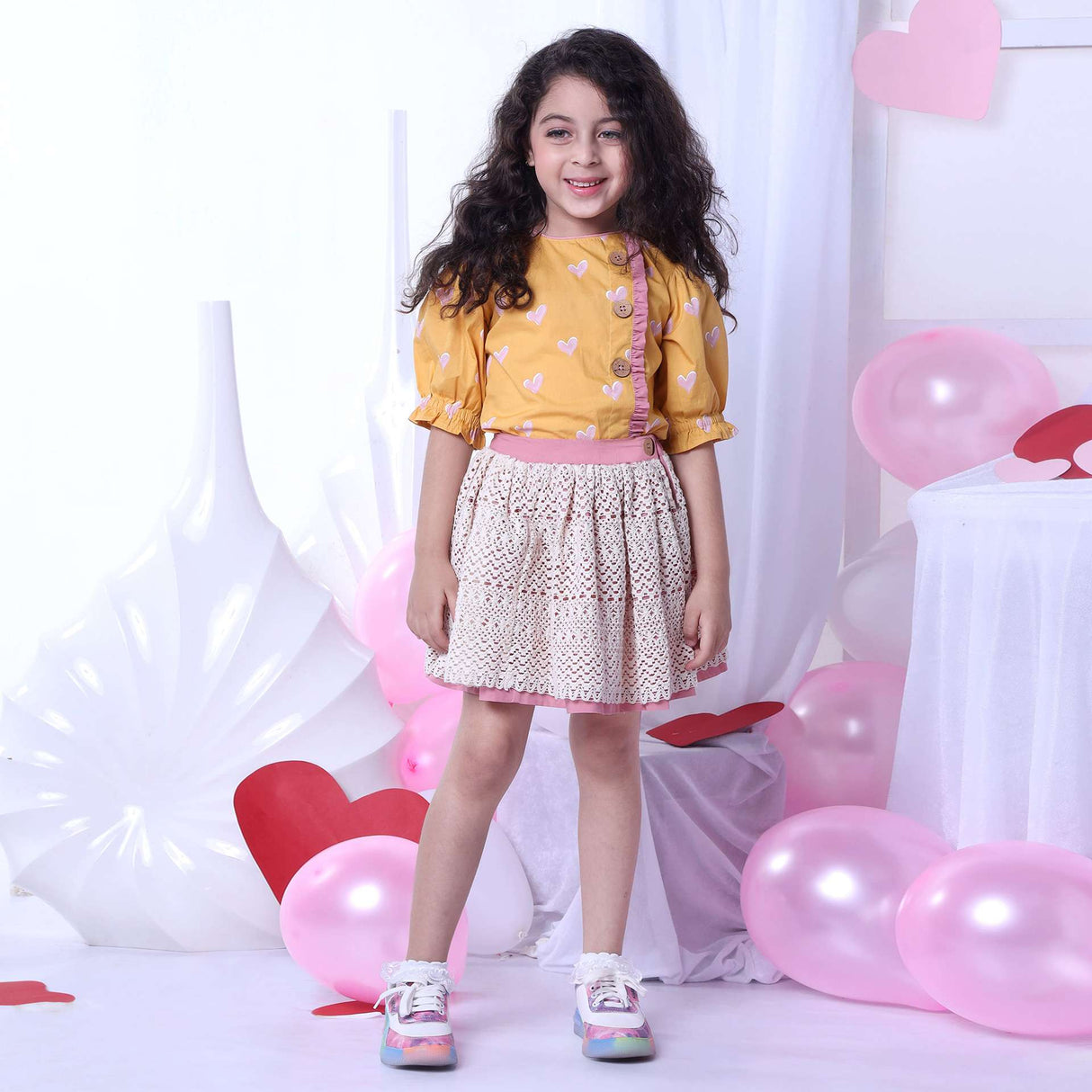 A girl in blouse with a heart print on a wheat gold base and gathered sleeves and ivory lace skirt with pink underskirt