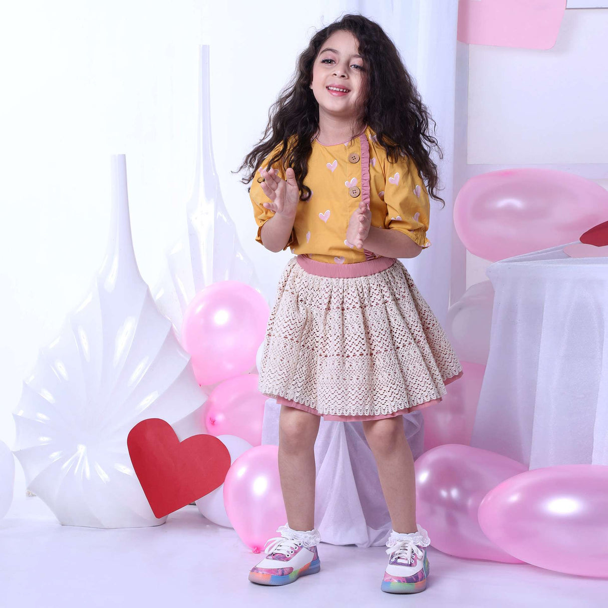 A girl in blouse with a heart print on a wheat gold base and gathered sleeves and ivory lace skirt with pink underskirt