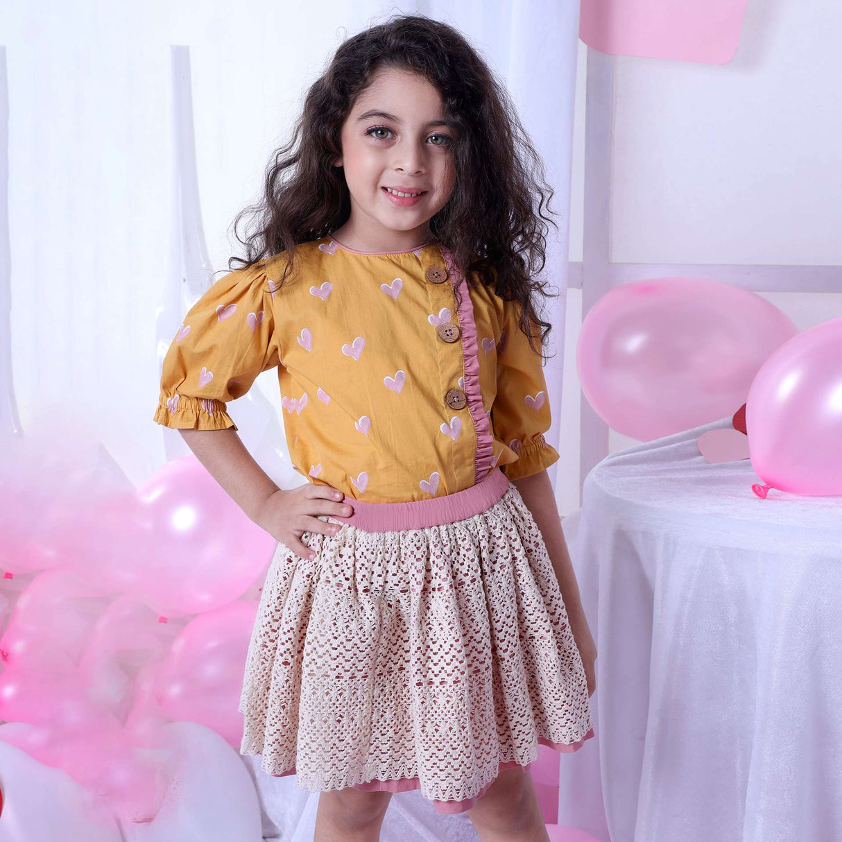 A girl in blouse with a heart print on a wheat gold base and gathered sleeves and ivory lace skirt with pink underskirt