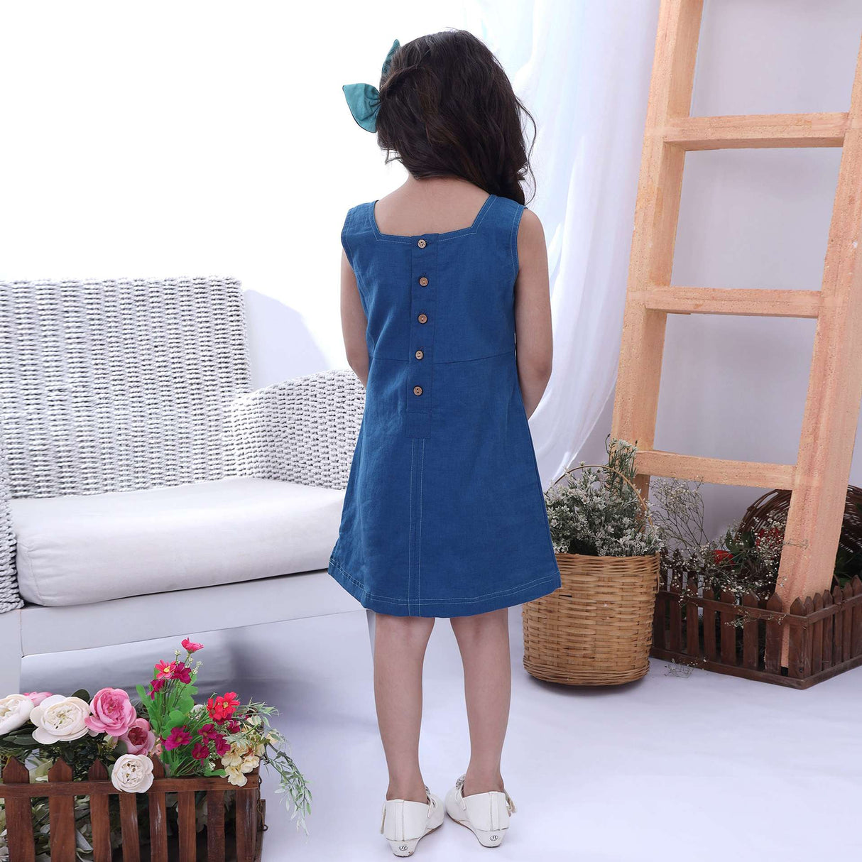 Rear image of a girl in a blue sleeveless dress with a hand embroidered bird cage and some scattered flowers