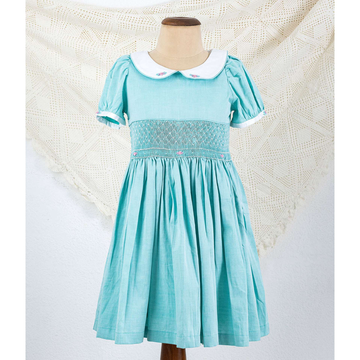 Mannequin has a turquoise classic smocked girls dress with white smocking and pink roses. The collar has embroidery roses and sleeves have white cuffs