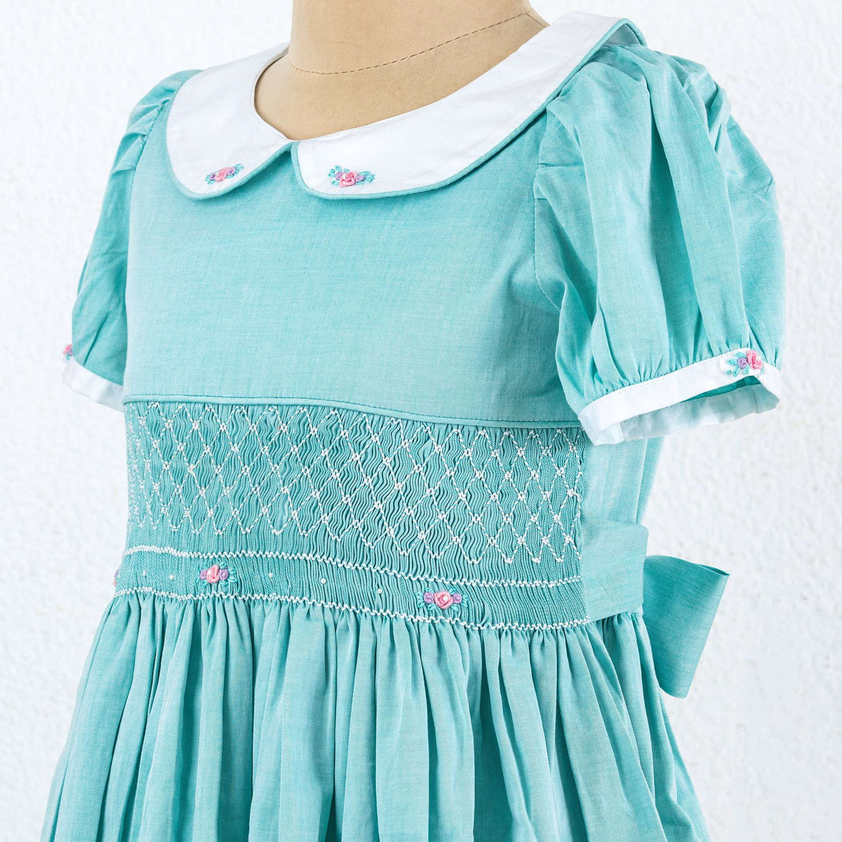 Turquoise girls dress on a mannequin with smocked band and a white collar and cuffs with small embroidered pink roses