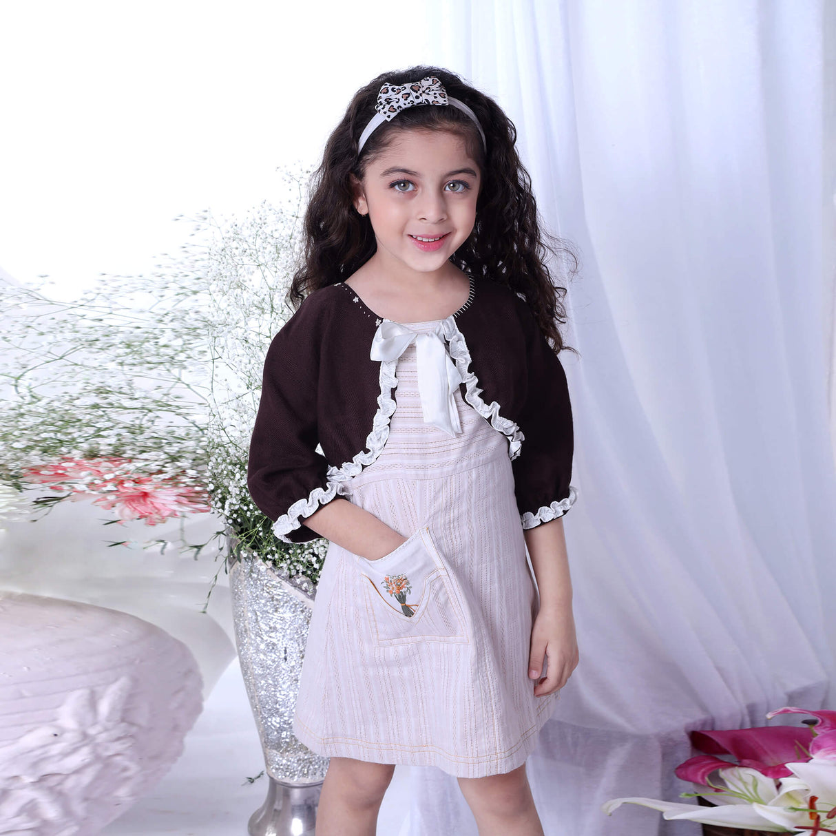 A girl in a ivory A-line dress paired with a brown bolero jacket, with her hand in an embroidered pocket