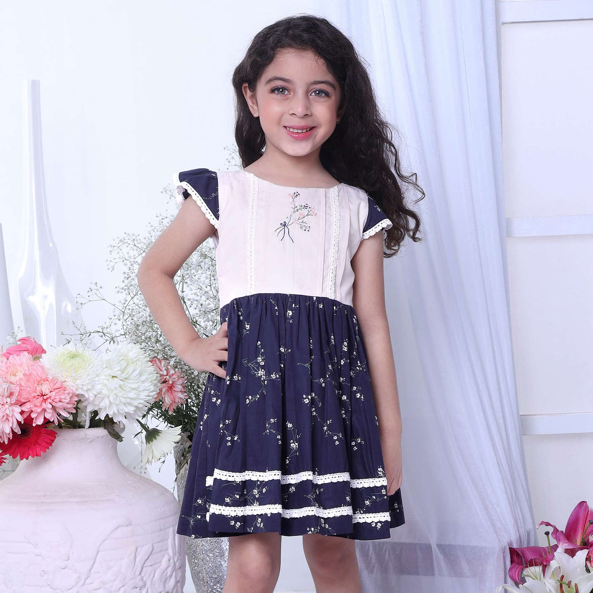 A girl in floral printed cotton navy dress with flutter sleeves, lace trims and hand embroidered bouquet in the yoke