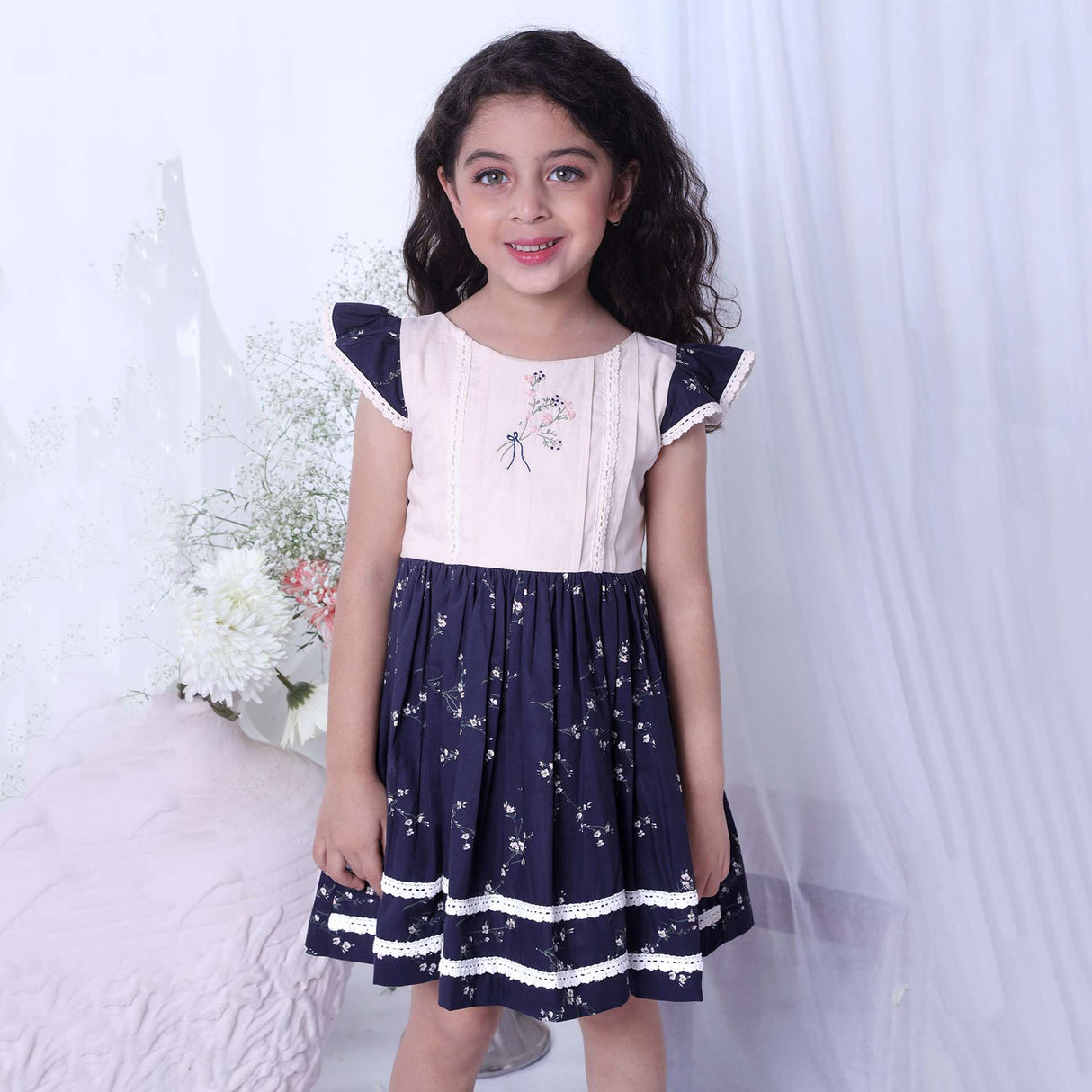 A girl in floral printed cotton navy dress with flutter sleeves, lace trims and hand embroidered bouquet in the yoke