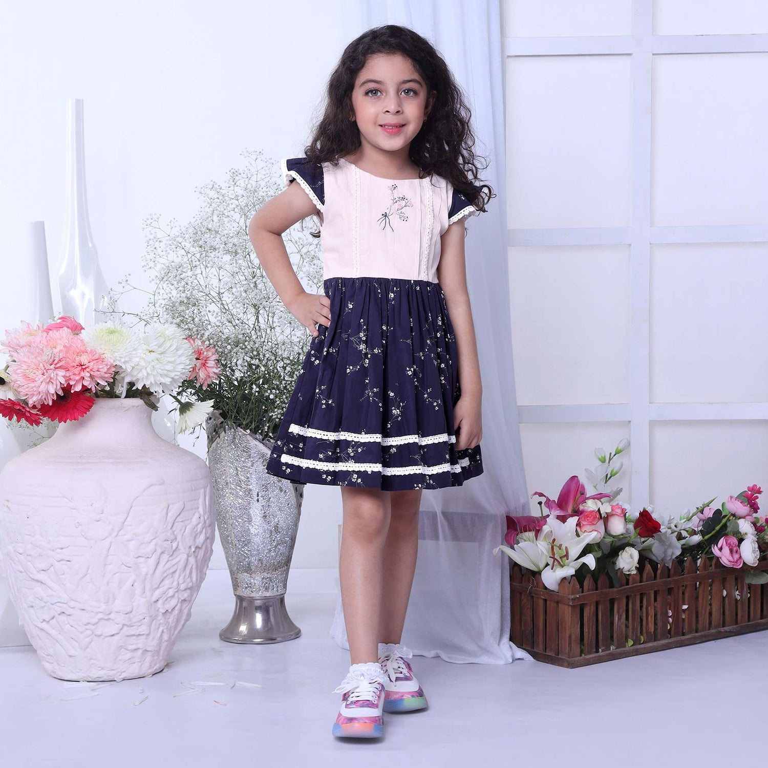 A girl in floral printed cotton navy dress with flutter sleeves, lace trims and hand embroidered bouquet in the yoke