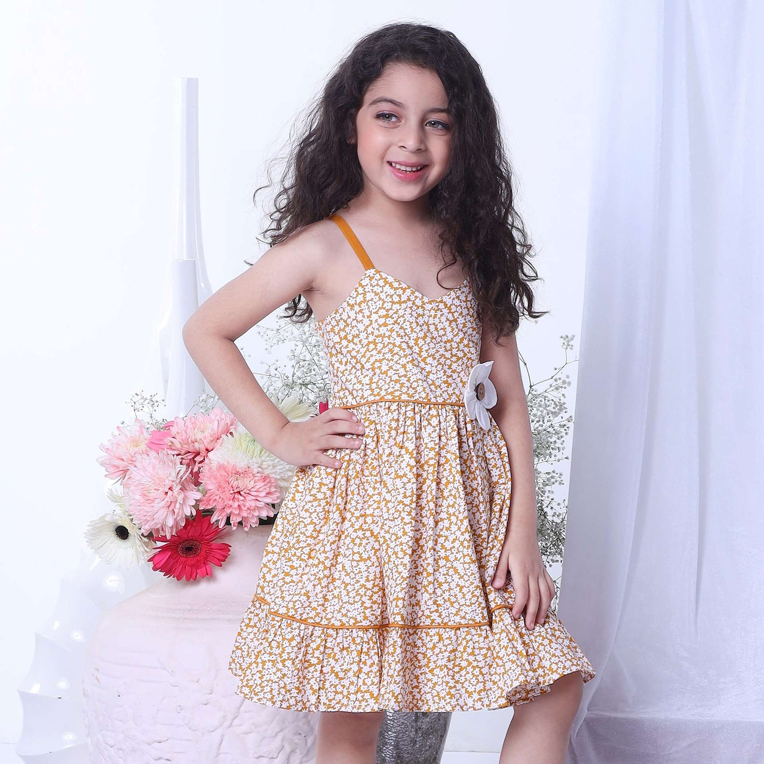 A little girl wearing strappy shoulder dress with floral print in mustard and big flower appliqued on the waist