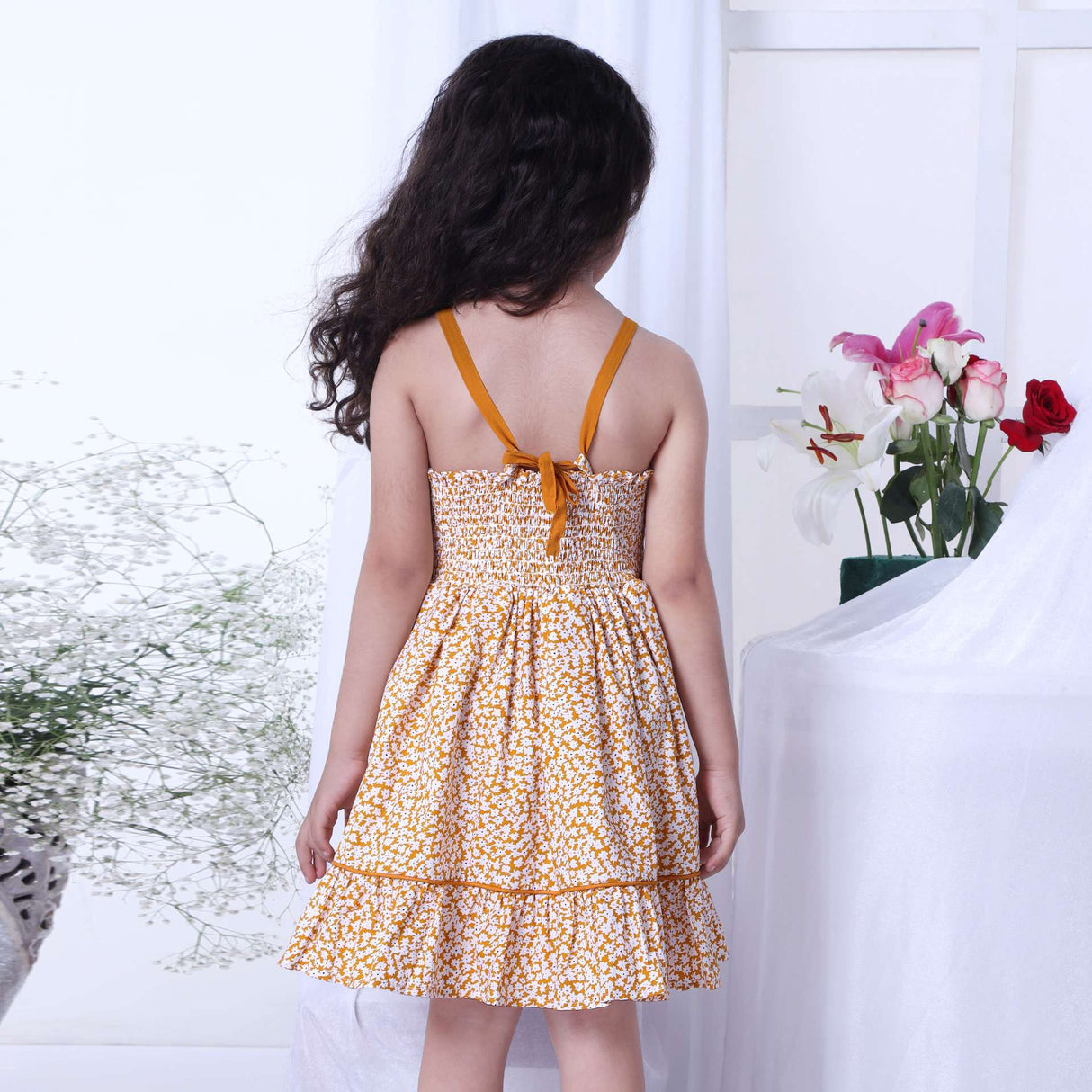A little girl wearing strappy shoulder dress with floral print in mustard and big flower appliqued on the waist