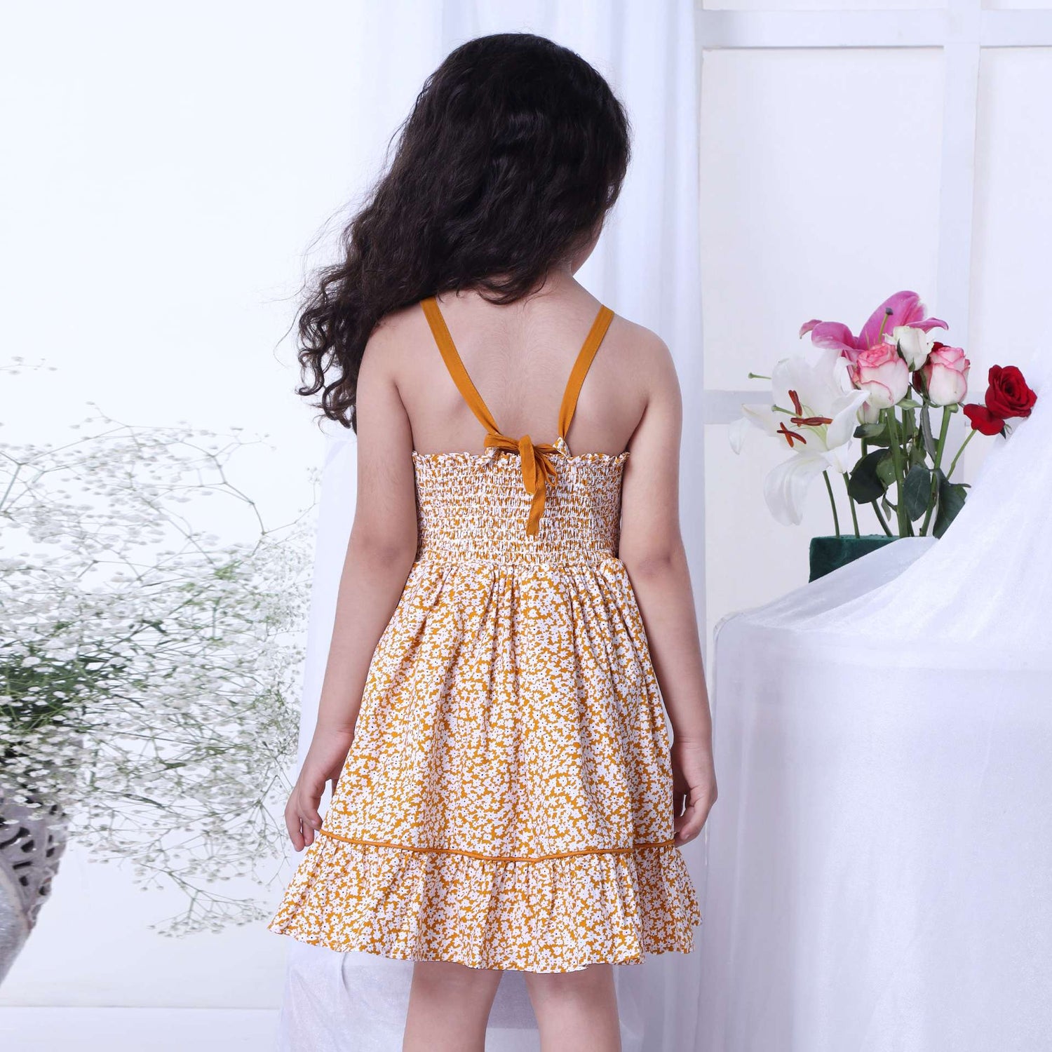A little girl wearing strappy shoulder dress with floral print in mustard and big flower appliqued on the waist