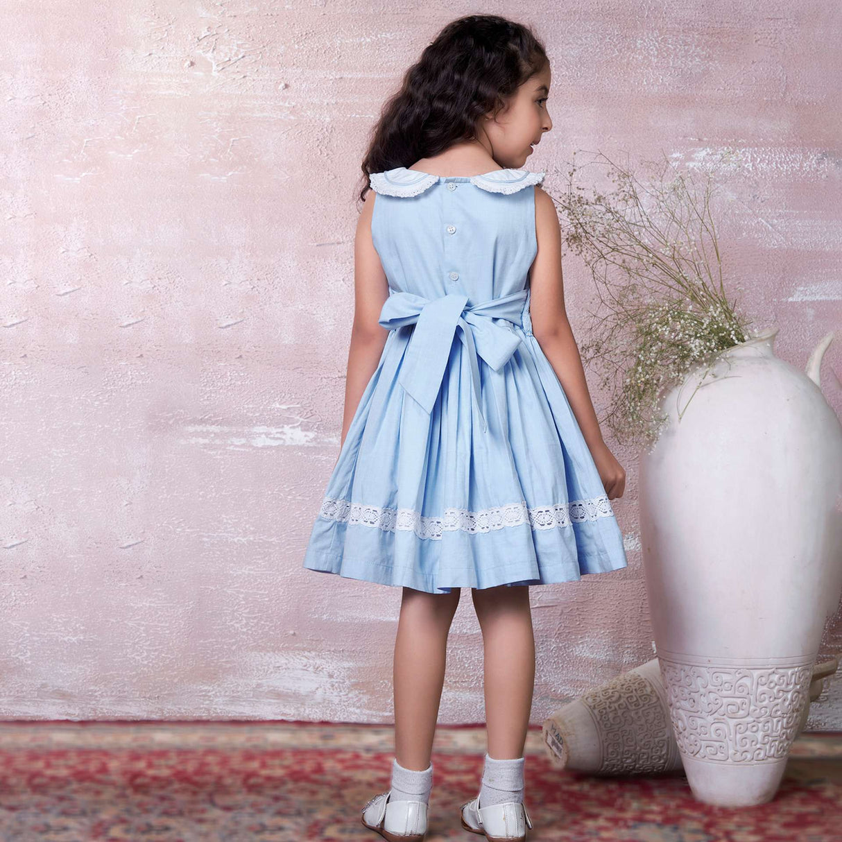 A little girl in blue hand-smocked dress with a lace insert on the skirt and a detailed collar with lace and cord piping