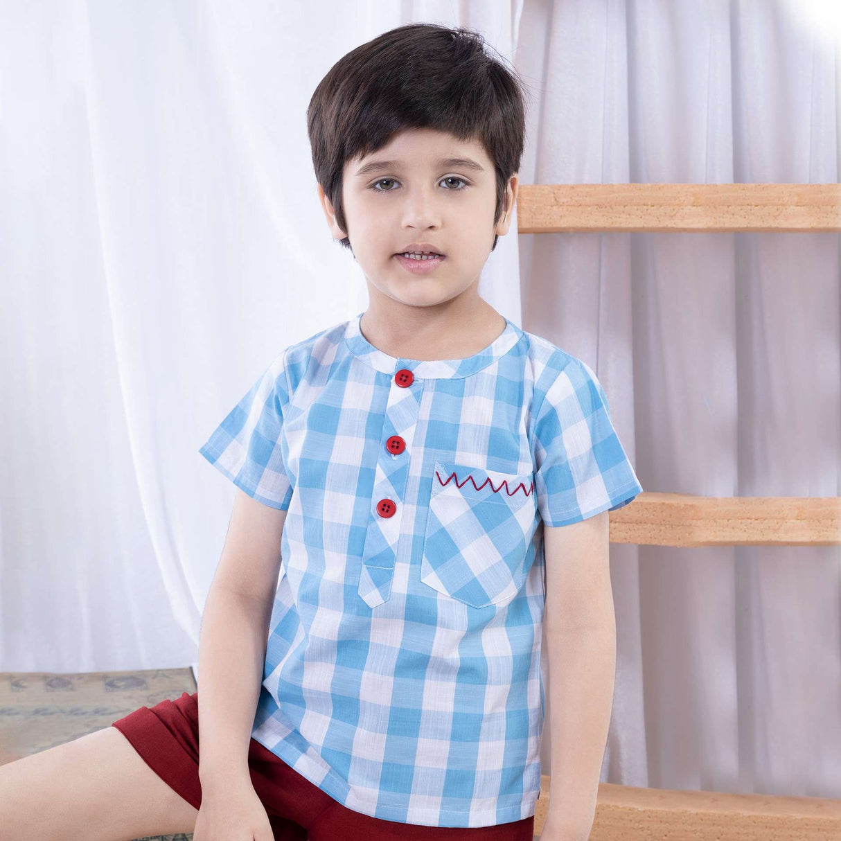 A little boy in light blue checked shirt with red hand embroidered pocket and red buttons paired with matching shorts