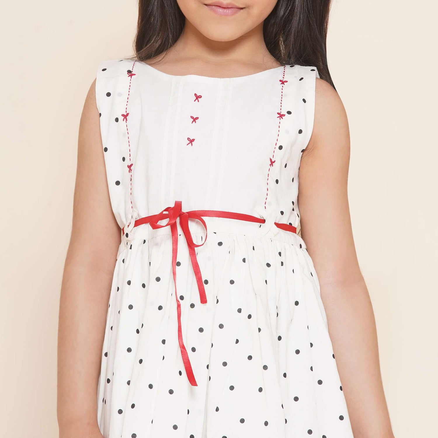 A little girl in sleeveless polka dot ivory cotton dress with red satin belt and hand embroidered bow embellishments