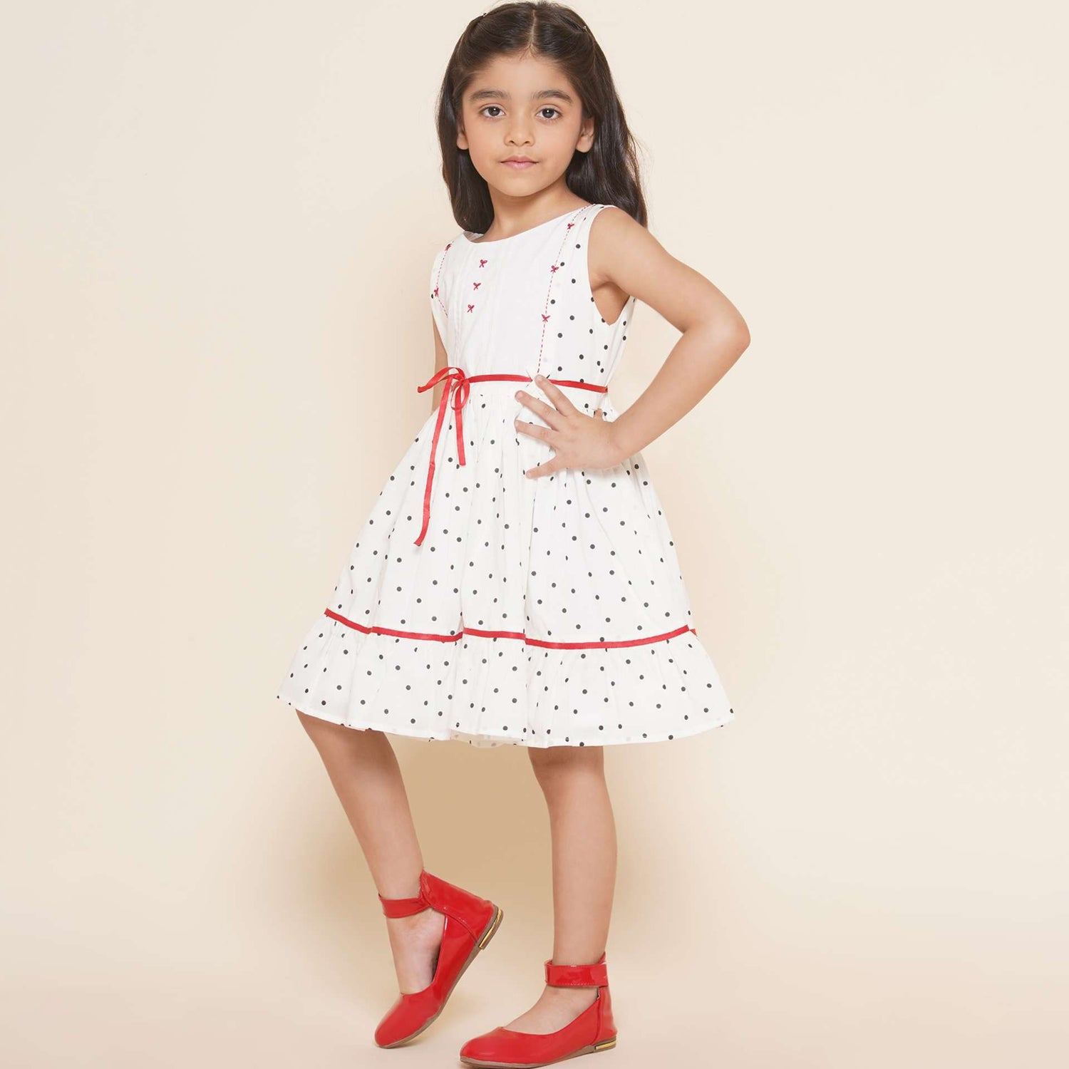 A little girl in sleeveless polka dot ivory cotton dress with red satin belt and hand embroidered bow embellishments