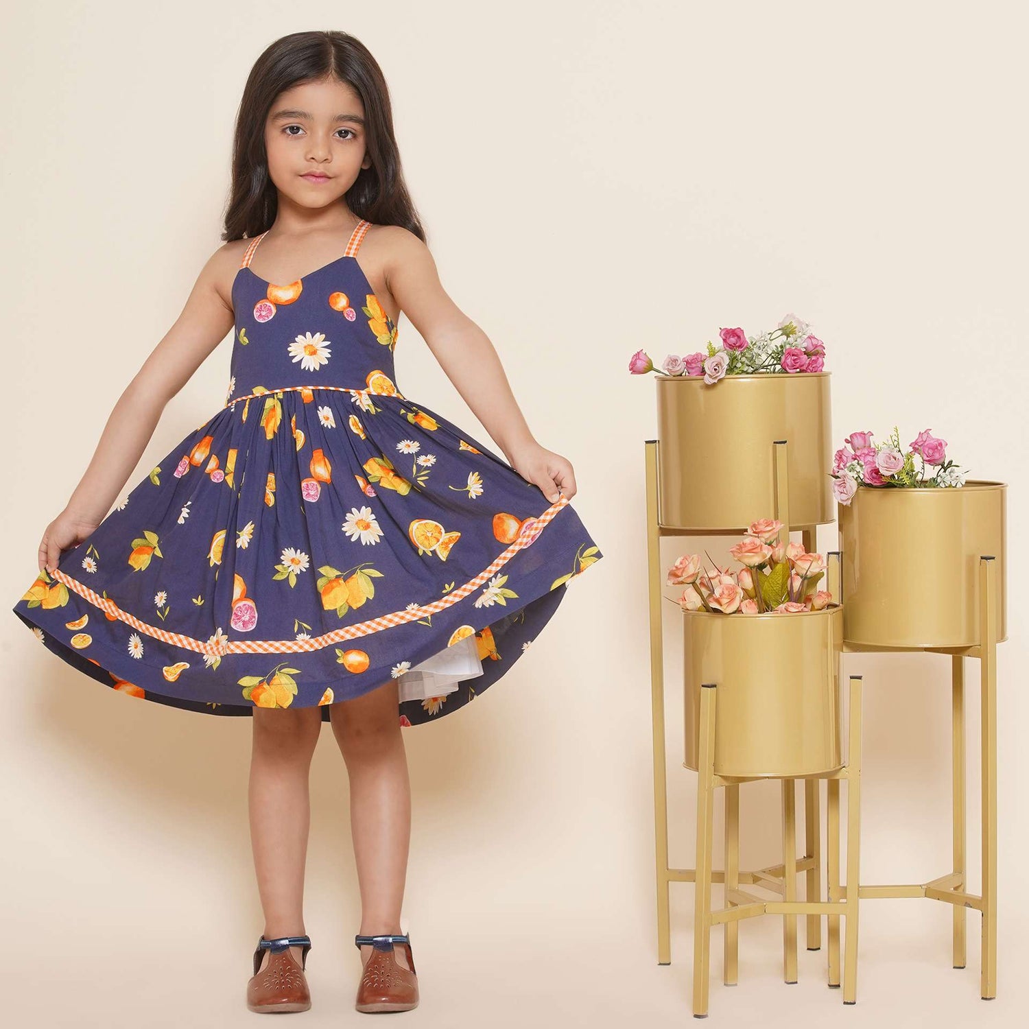 A little girl wearing strappy vibrant fruit printed dress with orange gingham trims, shirred back and tie-up straps