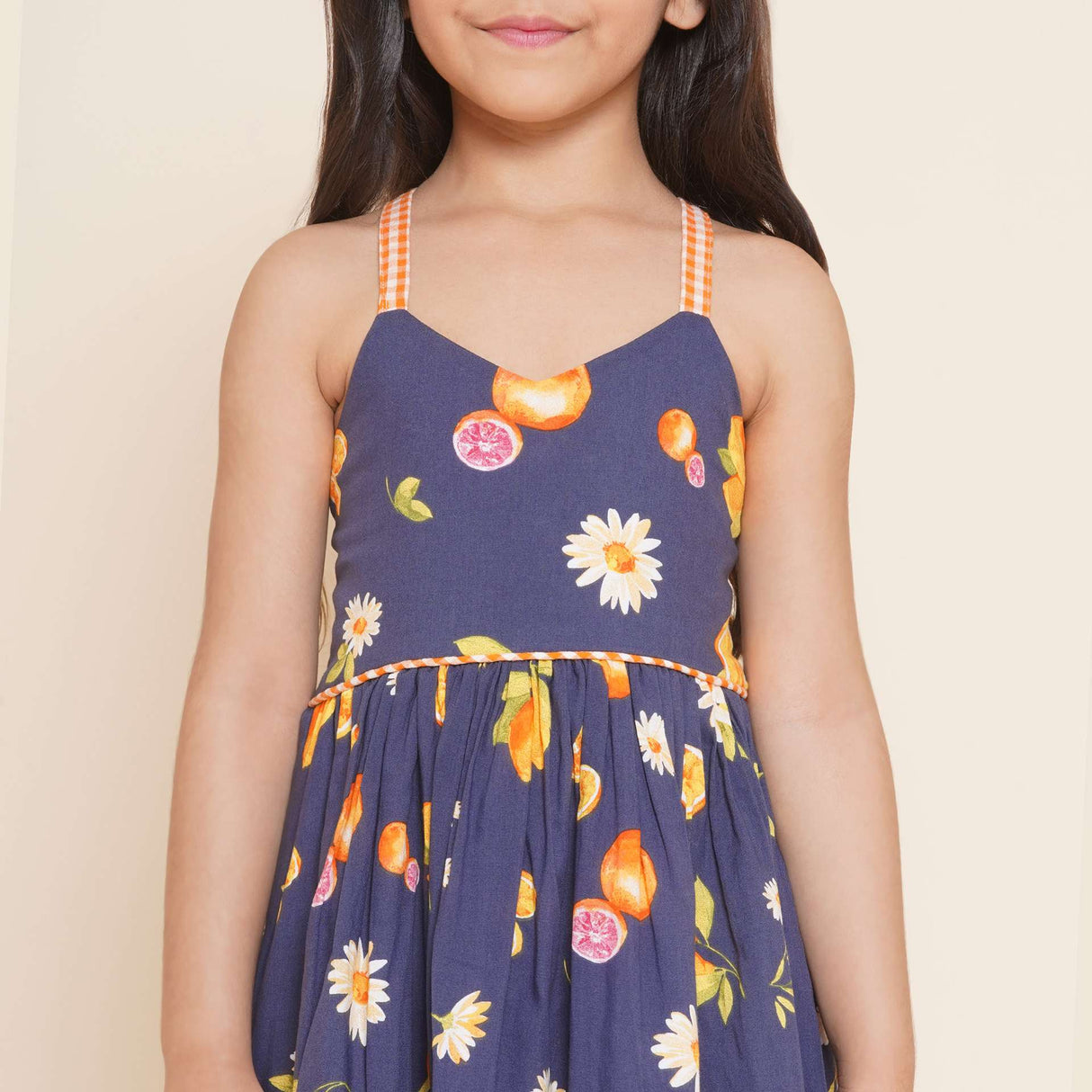 A little girl wearing strappy vibrant fruit printed dress with orange gingham trims, shirred back and tie-up straps