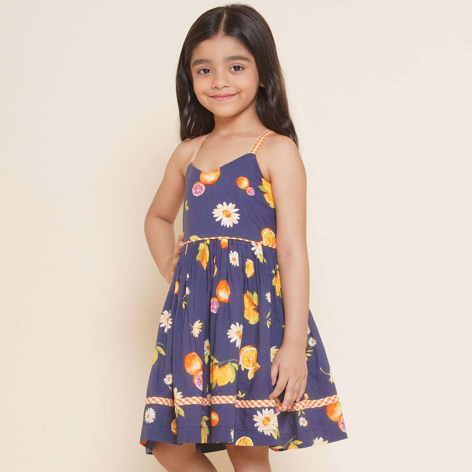 A little girl wearing strappy vibrant fruit printed dress with orange gingham trims, shirred back and tie-up straps