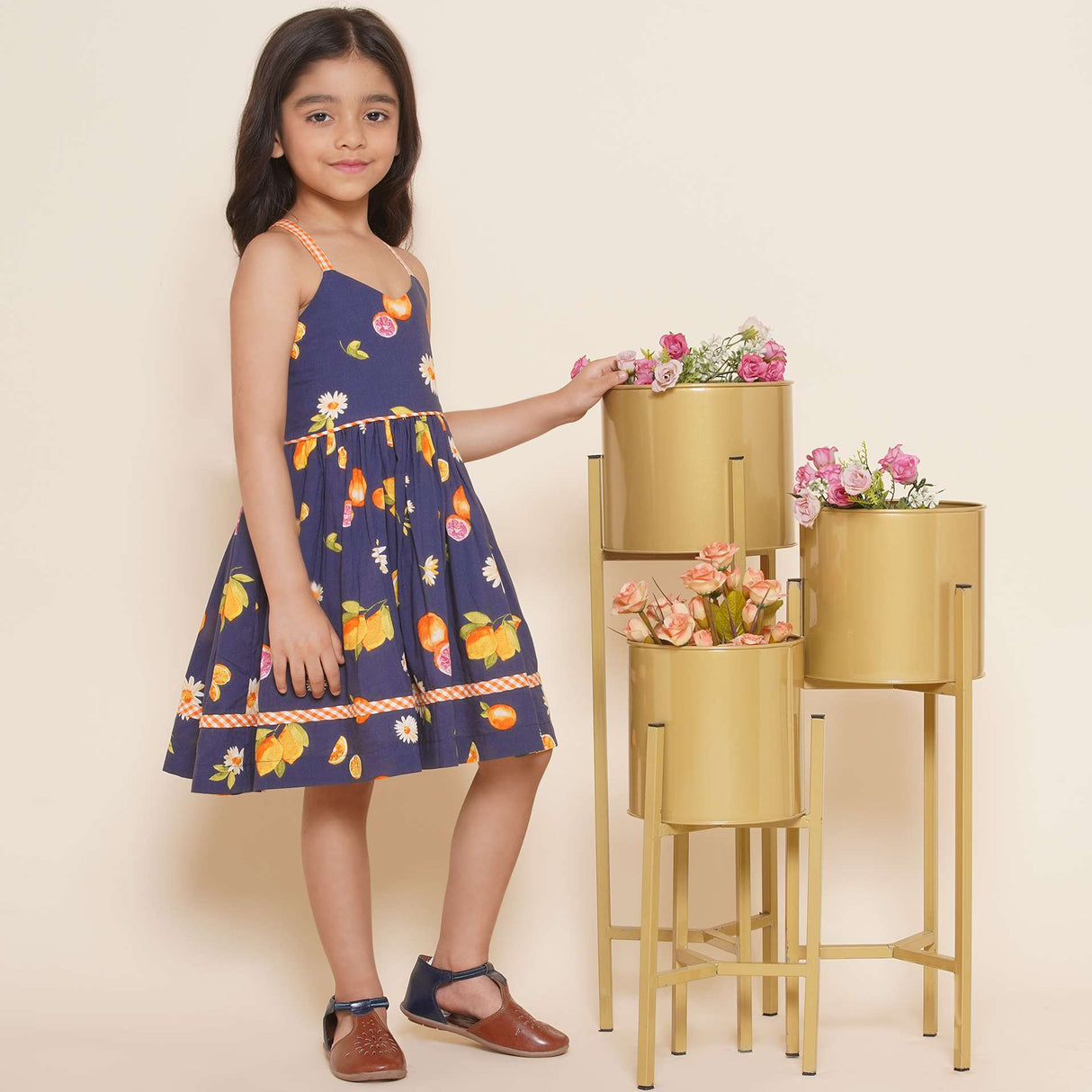 A little girl wearing strappy vibrant fruit printed dress with orange gingham trims, shirred back and tie-up straps