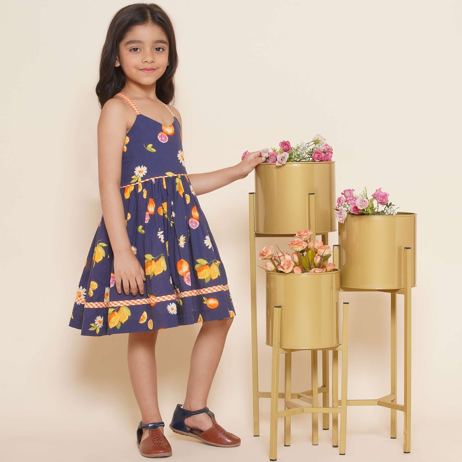 A little girl wearing strappy vibrant fruit printed dress with orange gingham trims, shirred back and tie-up straps