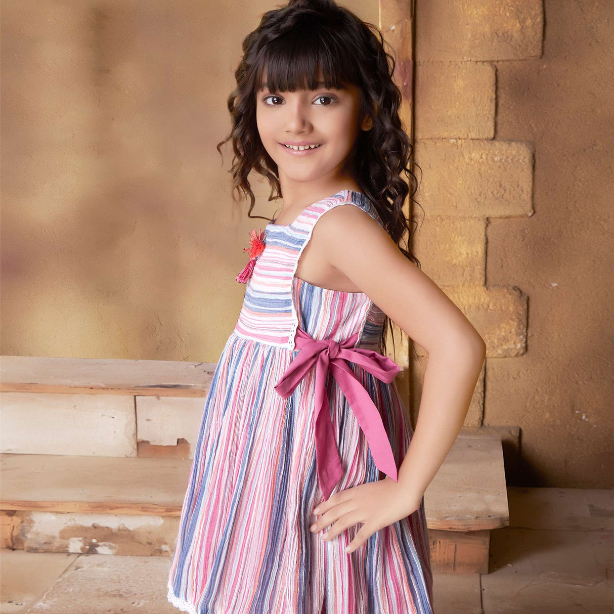 A little girl in sleeveless multi-color stripe dress with side ties, lace trims and a decorative thread bow