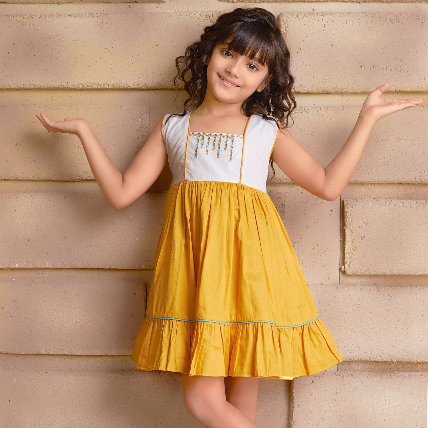 A young girl wearing sleeveless golden yellow dress with a 'Toran' hand embroidered yoke and hem ruffles