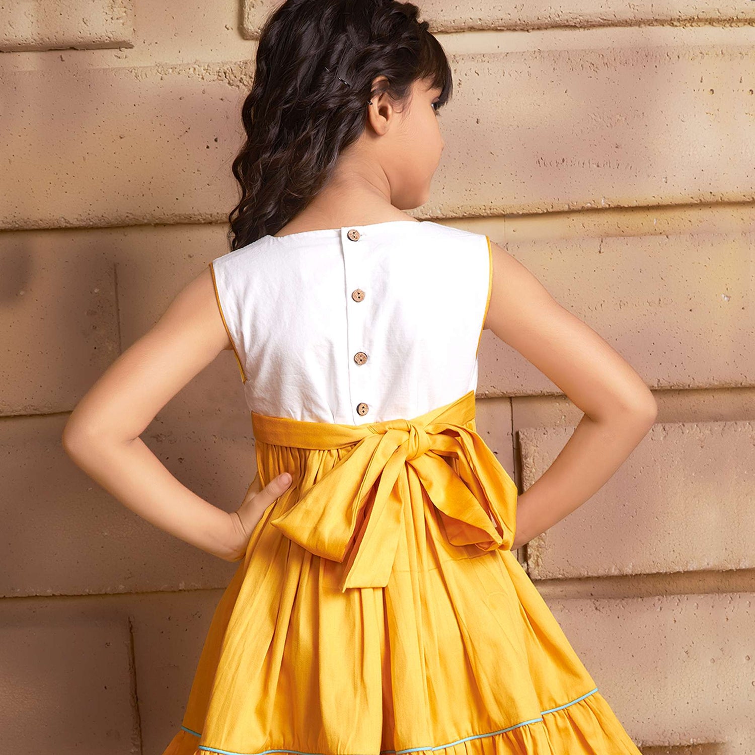 Rear image of a girl wearing sleeveless golden yellow dress with a 'Toran' hand embroidered yoke and hem ruffles