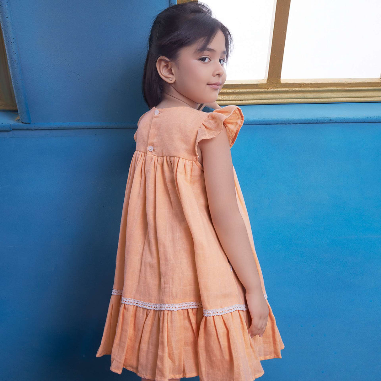 Rear image of a girl in peach gauze dress with hand-embroidered flowers in the yoke, ivory lace trims and a ruffled hem