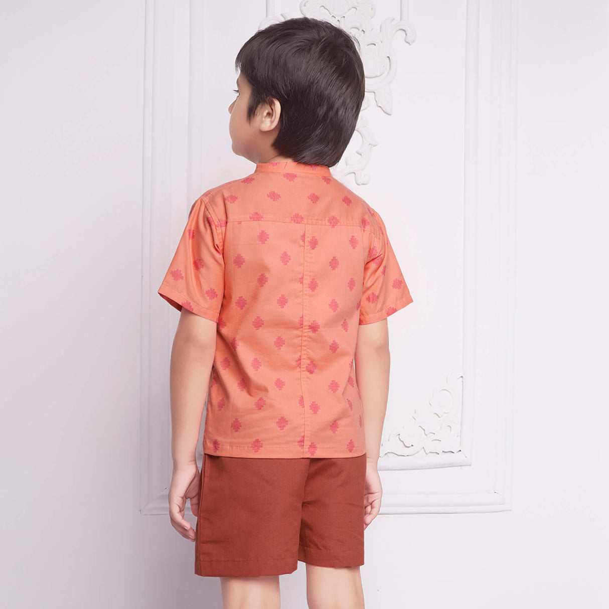Back view of a boy wears peach printed shirt matched with rust shorts, both with pockets.