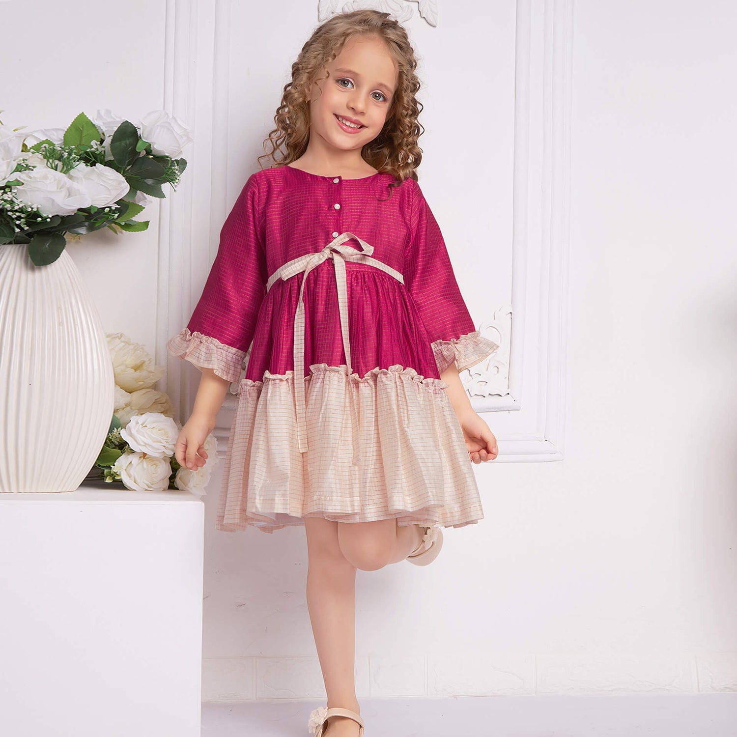 A girl wearing a pink and ivory Chanderi full sleeve ruffle dress, standing with straight posture