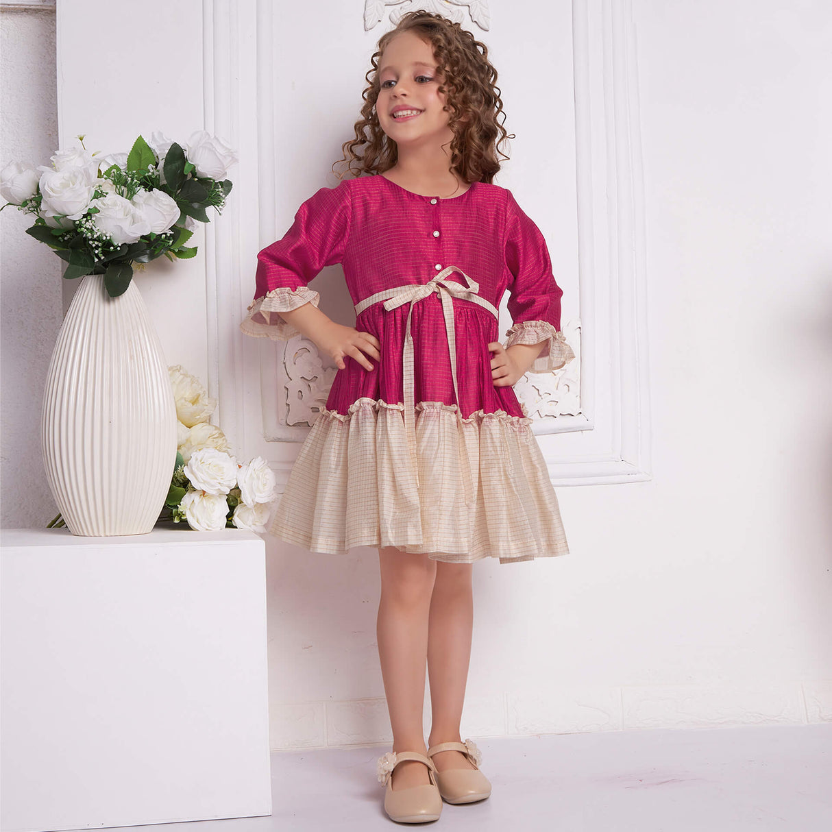 A girl poses in a pink and ivory Chanderi ruffle dress with hand on hips, featuring front pearl buttons and a tie-up sash