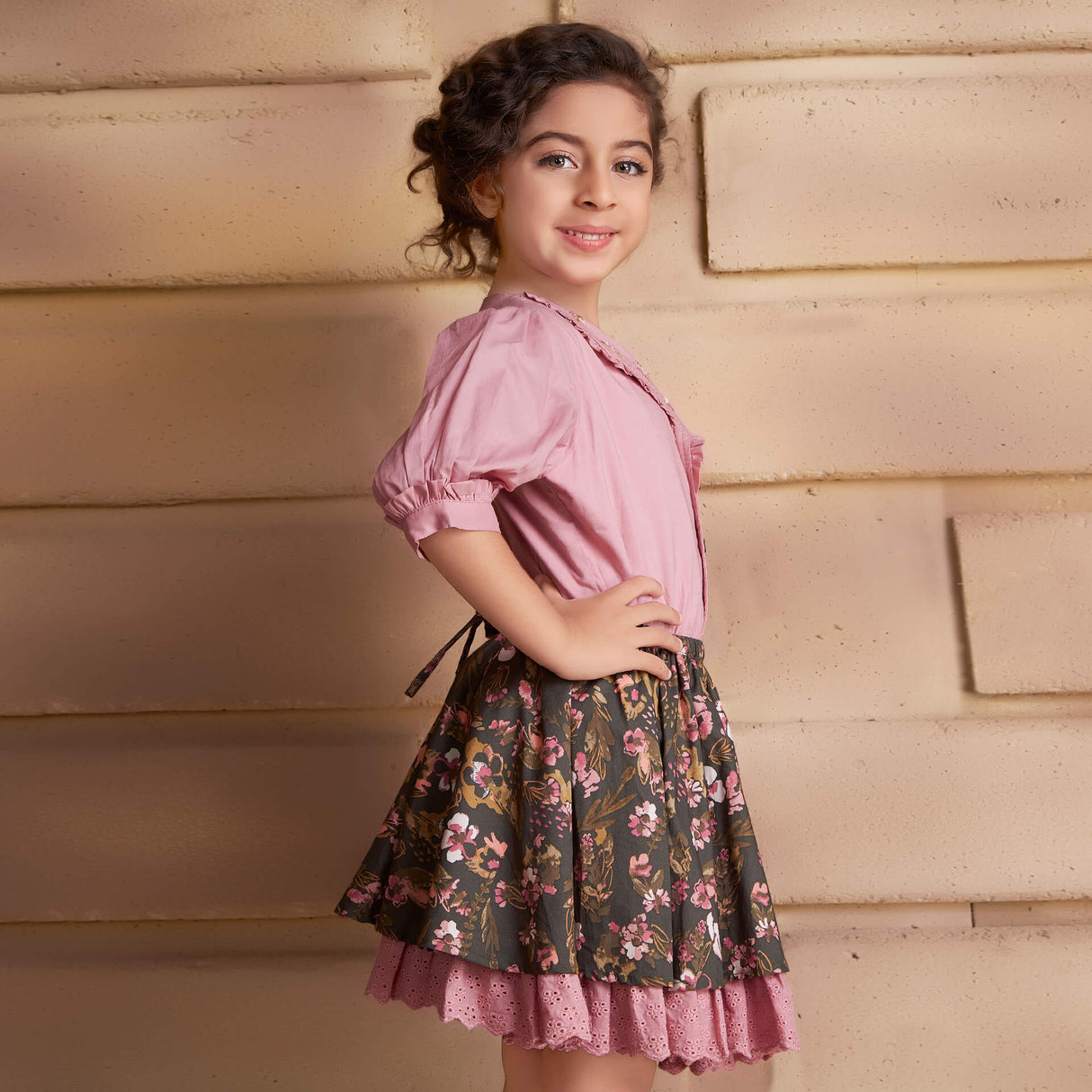 Posing from the side, a girl wears a pink embroidered blouse and floral printed skirt