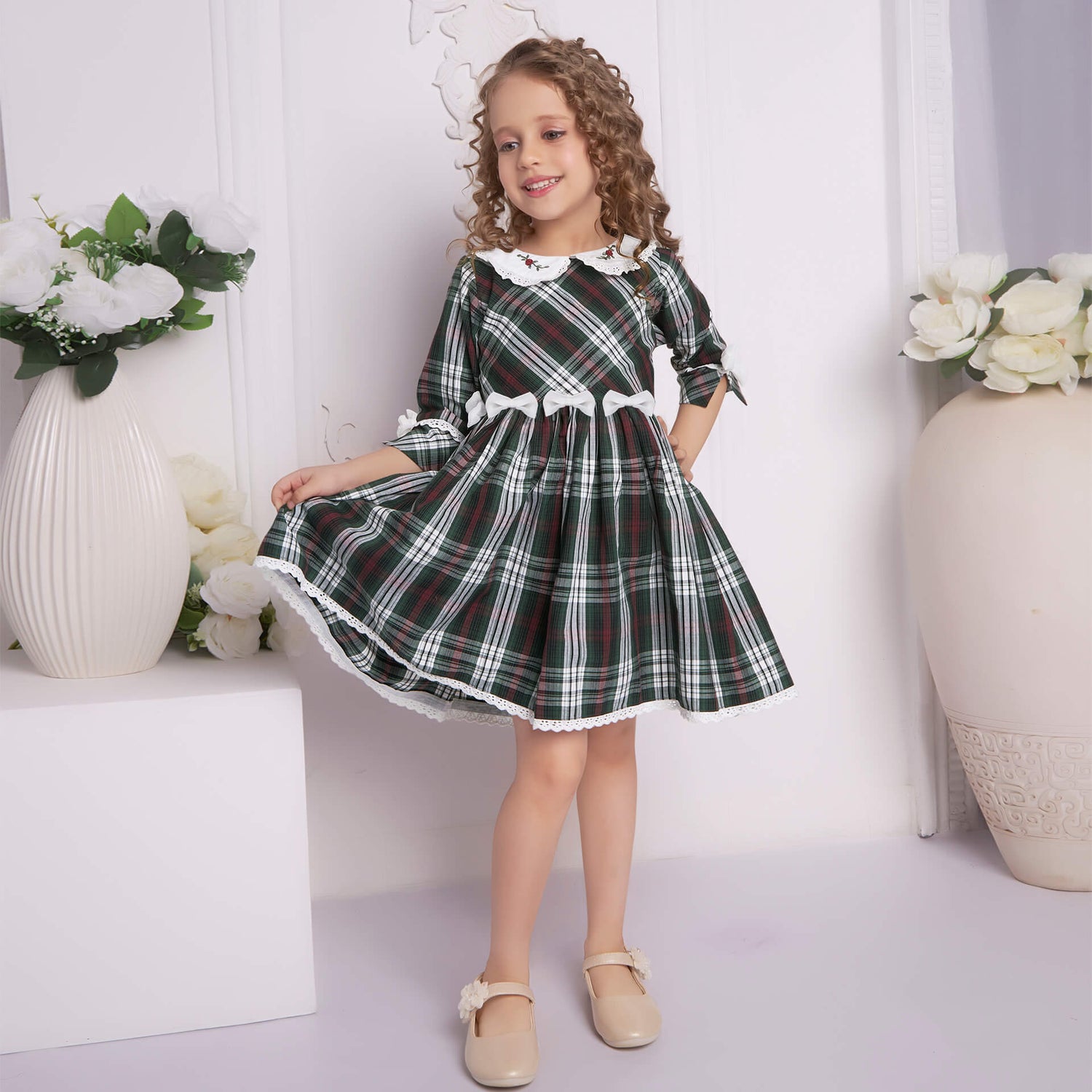 Elegantly posing, a young girl showcases a green tartan collar embroidered dress with bows at the waist