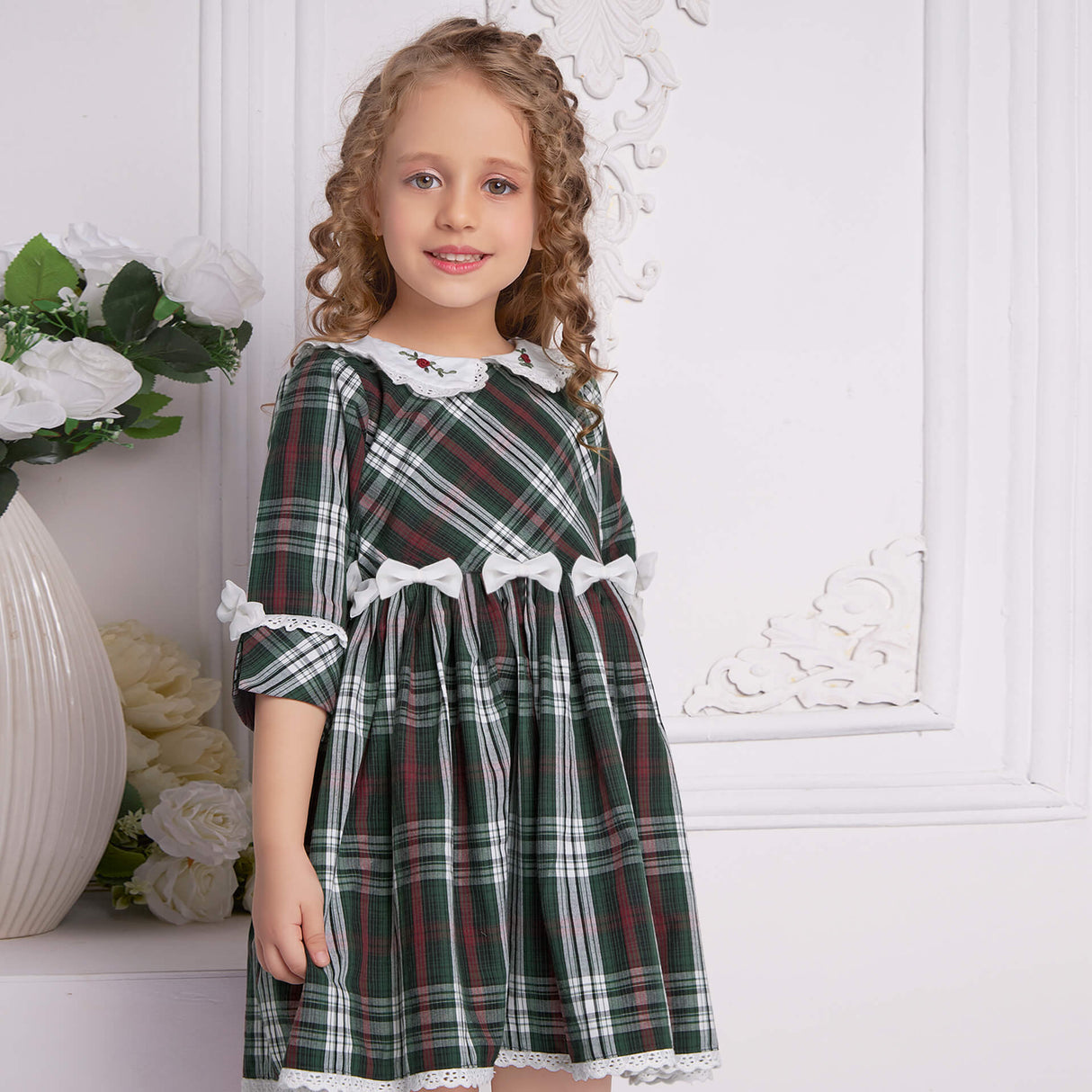 A young girl showcases a green tartan collar embroidered dress with bows at the waist in a slightly turned side view