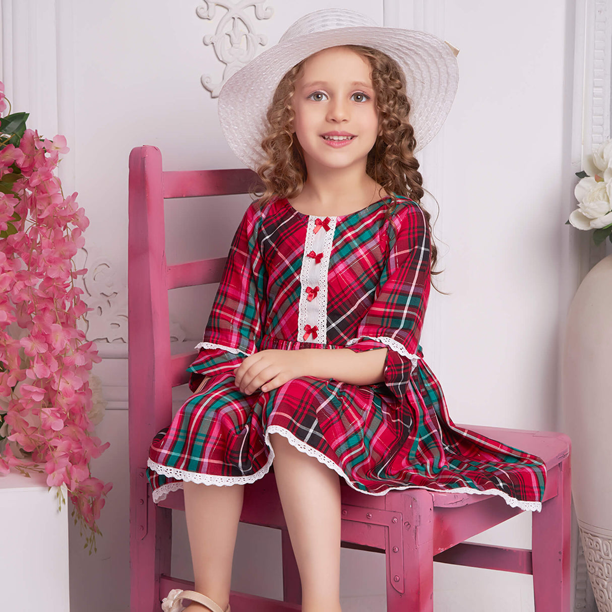 Seated with a hat atop her head, a little girl wears red plaid dress with white lace and red satin bows as trims