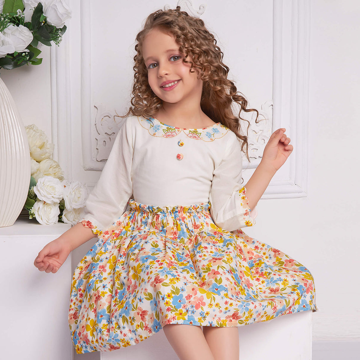 A little girl seated wearing ivory floral dress with delicate floral hand-embroidered neckline