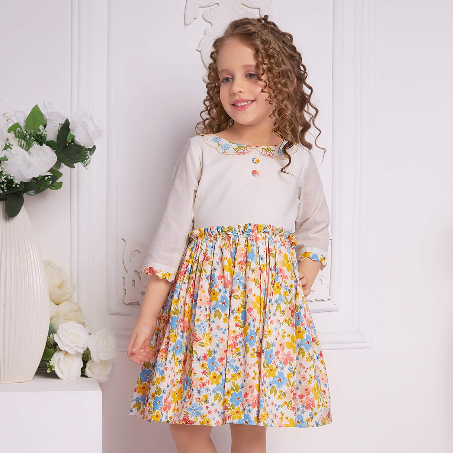 With hand on hips, a girl poses in an ivory floral embroidered dress with decorative cloth buttons