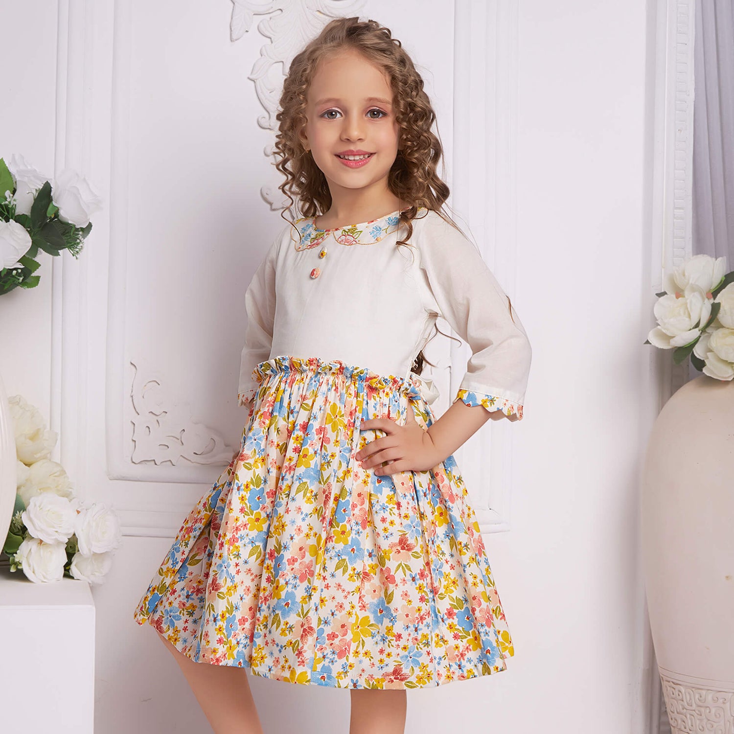With hands on hips, a side pose of a little girl in an ivory floral embroidered dress with decorative cloth buttons