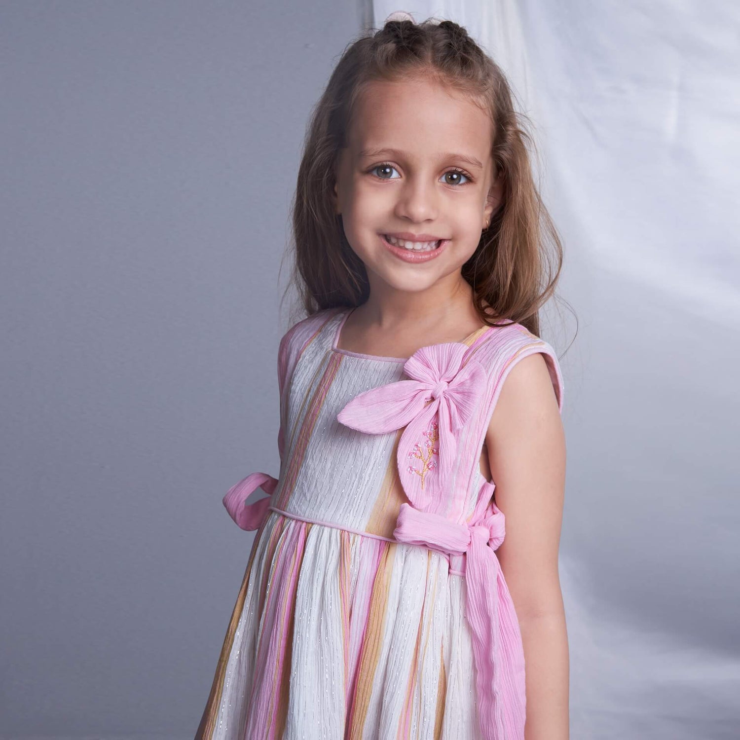 Side view close-up of a girl in a sleeveless pink lurex embroidered dress with side ties and an attached embroidered bow near the neckline.