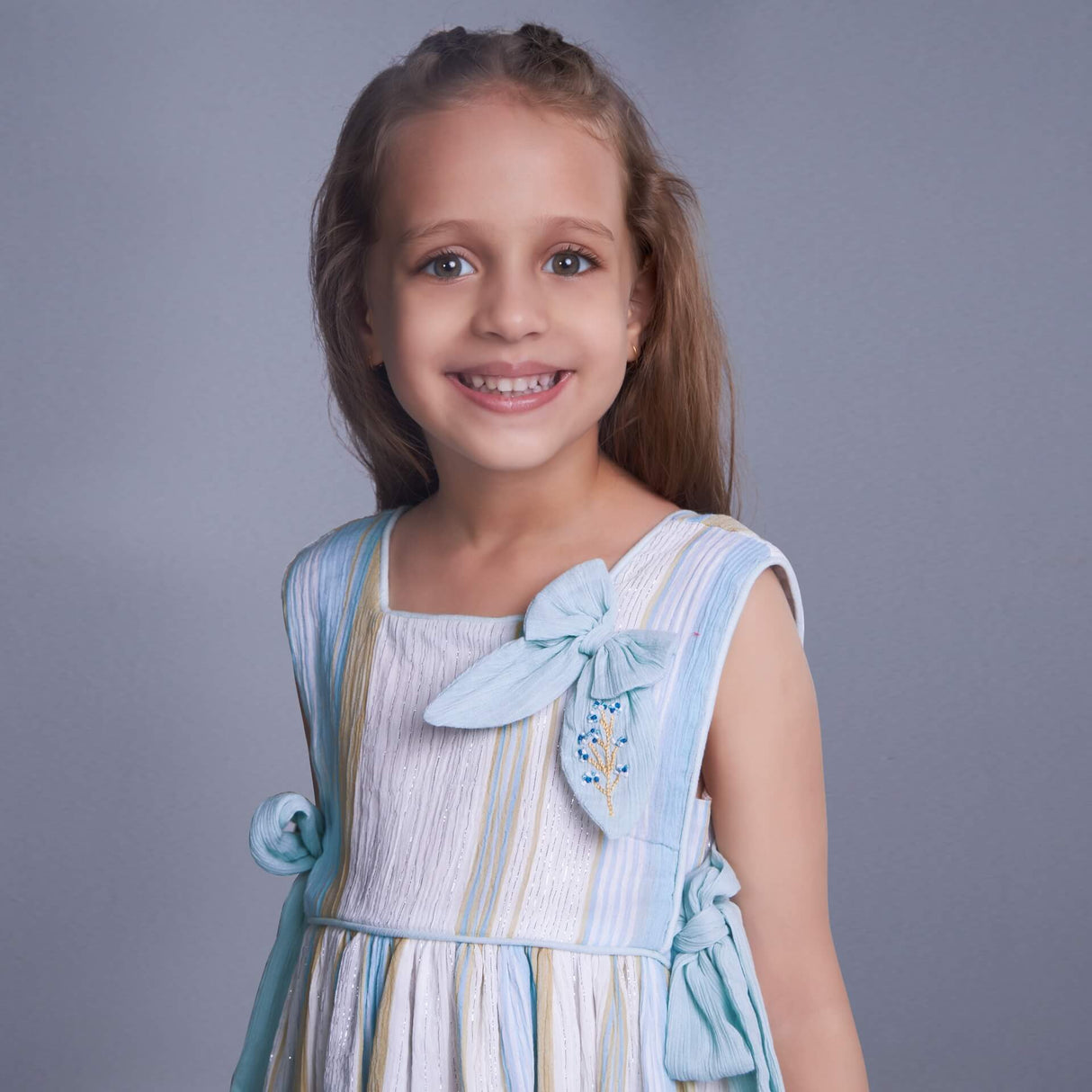 In a focused view, a girl is dressed in a blue lurex dress with an attached embroidered bow near the neckline