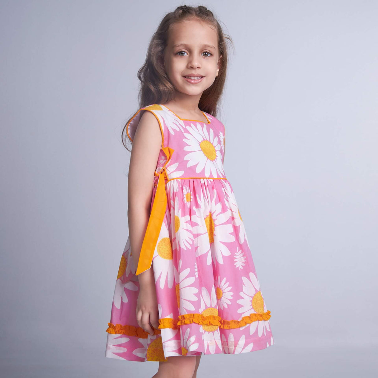 Posing from the side, a little one wears a pink floral dress with side ties in a contrasting tone