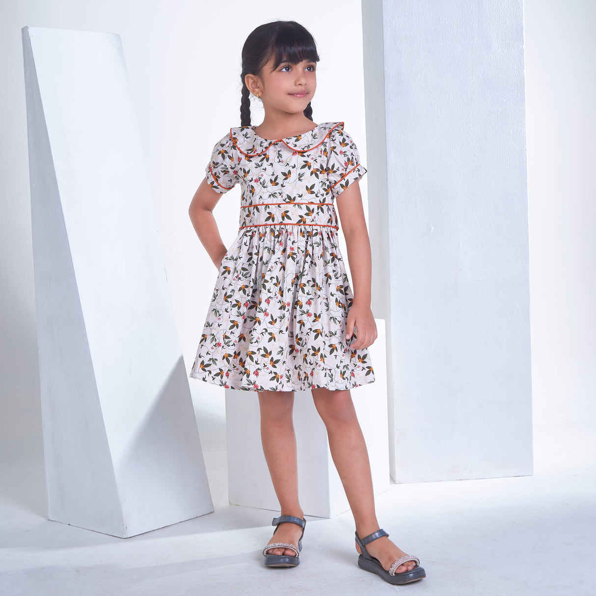 A girl stands in an ivory floral printed collared dress with an adjustable belt at the waist