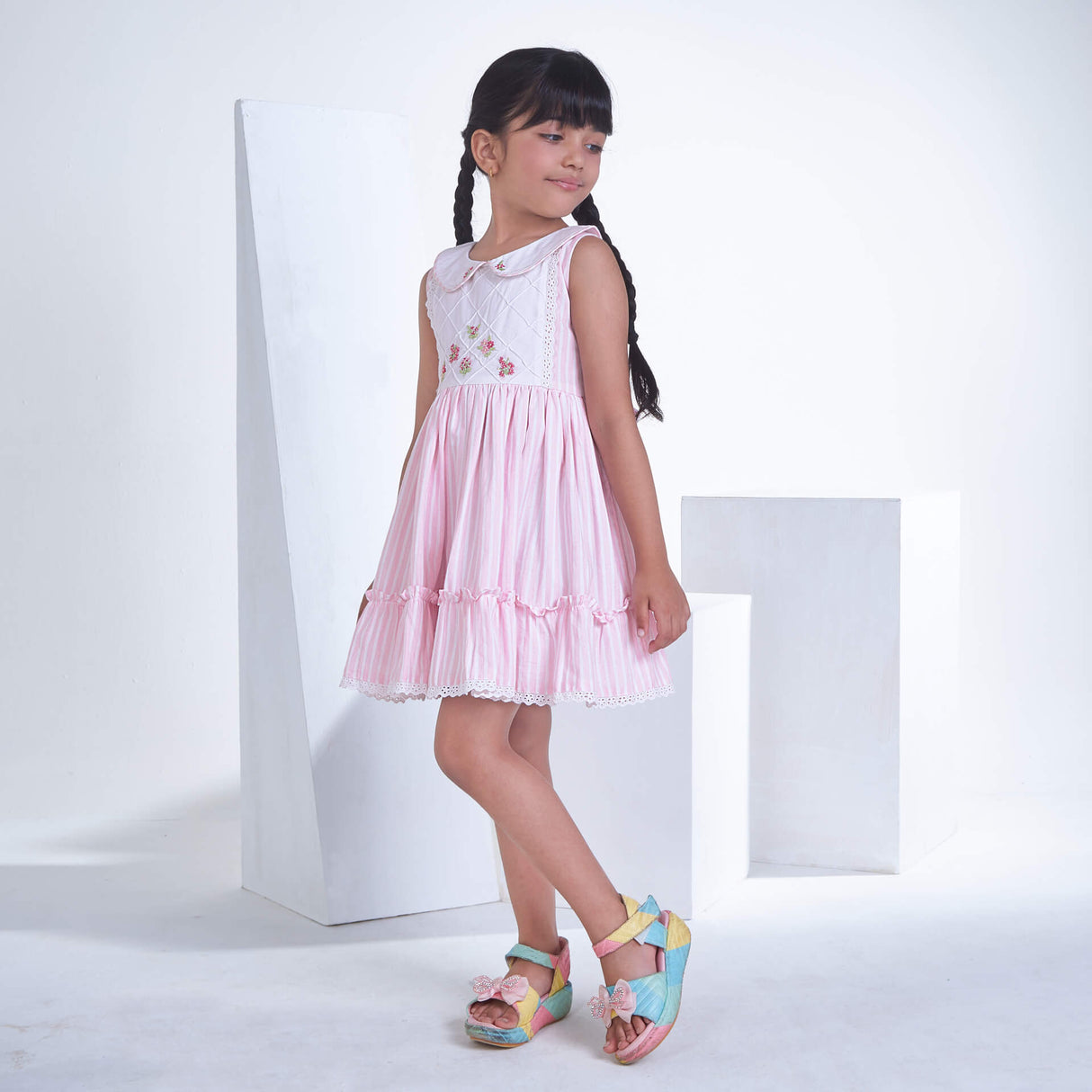 Posing from the side, a little one wears a pink striped embroidered collared dress adorned with ruffled hem