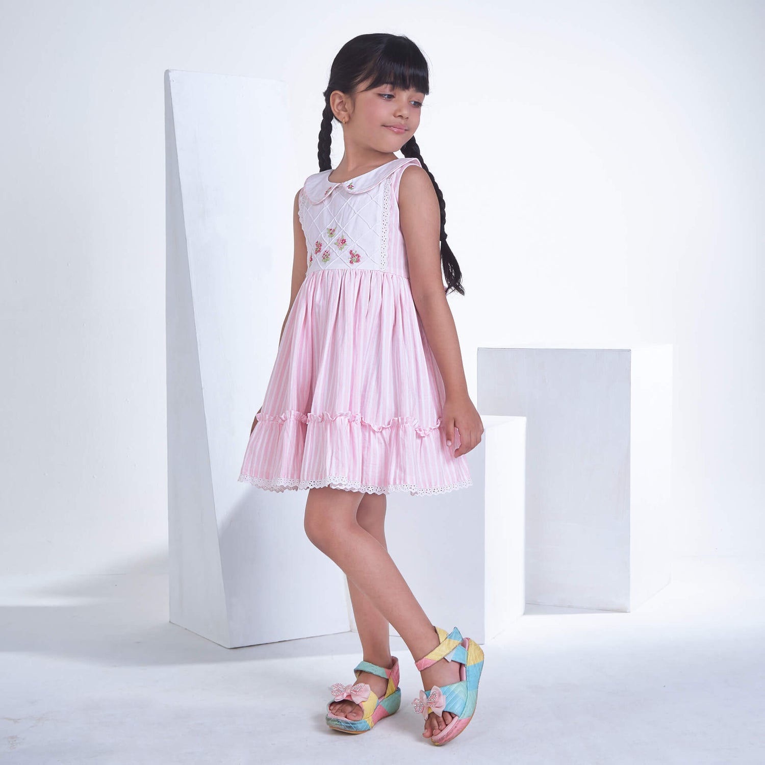 Posing from the side, a little one wears a pink striped embroidered collared dress adorned with ruffled hem
