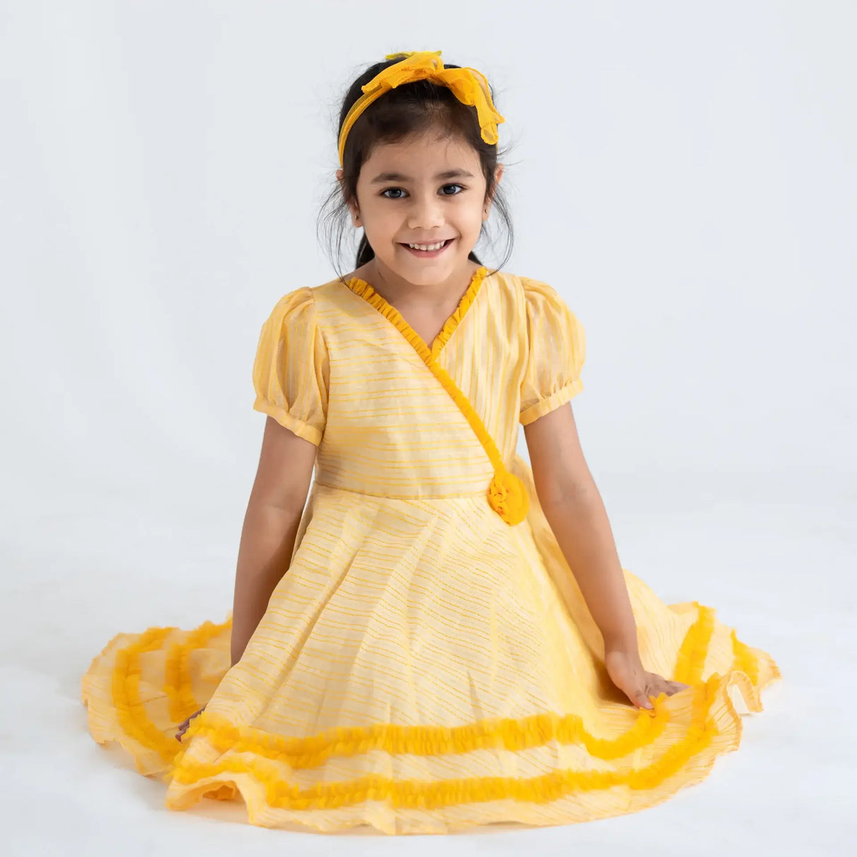 "A girl seated, wearing a yellow crossover chanderi puffed sleeves dress."