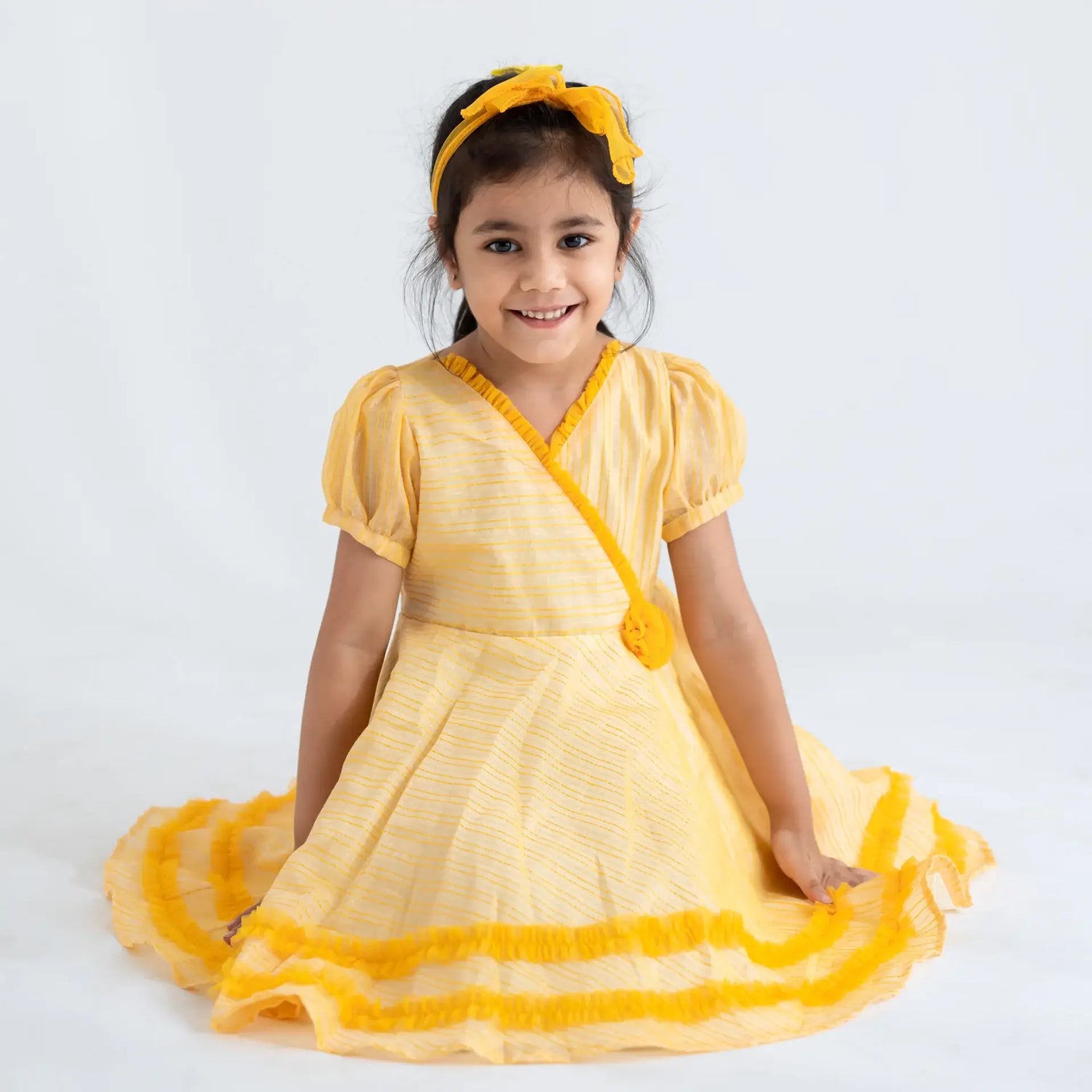 "A girl seated, wearing a yellow crossover chanderi puffed sleeves dress."